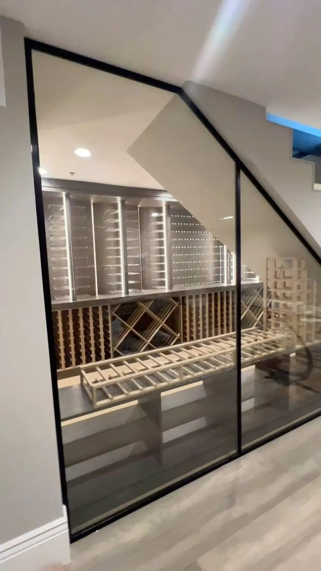Angled black-framed glass wall enclosing a wine room under stairs
